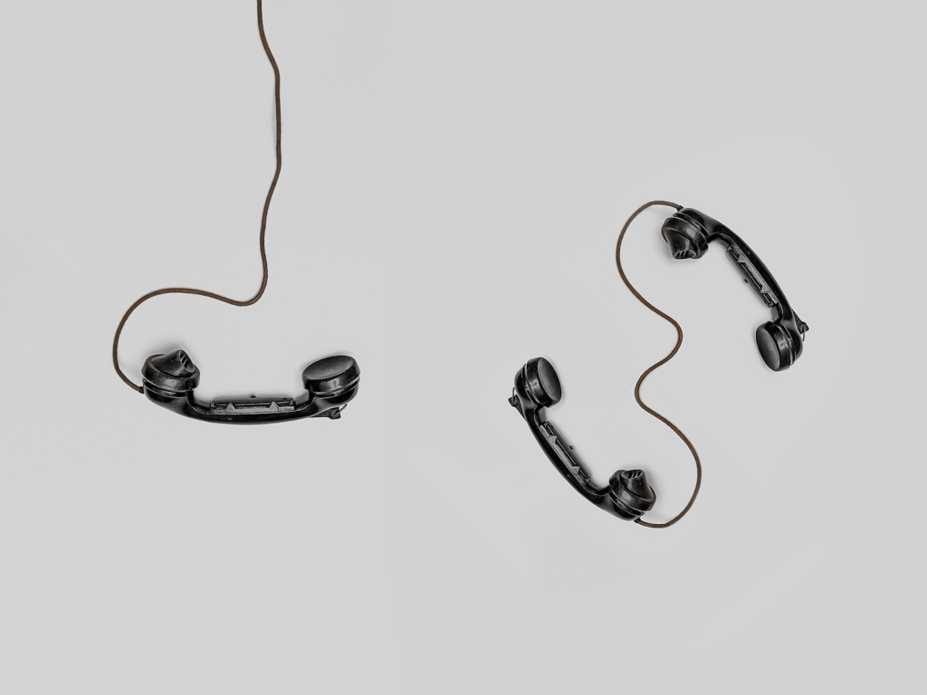 Two black vintage telephone receivers connected by a single cord on a plain grey background
